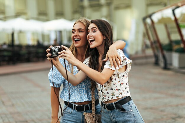 Charming brunette and blonde women in stylish floral blouses and denim pants smile and hug outside