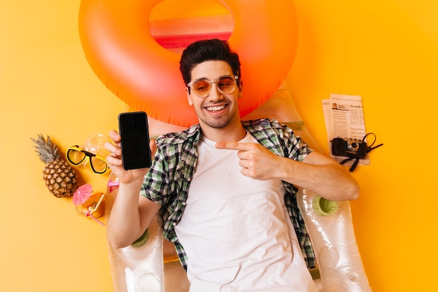 Free photo charming brunette man in plaid shirt and orange glasses points to black smartphone. guy is resting on inflatable mattress.