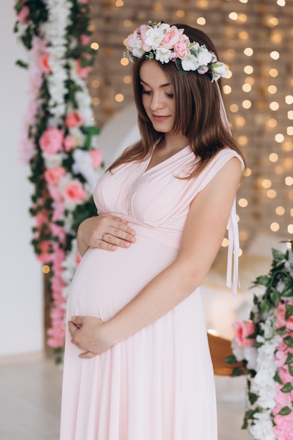 Charming brunette pregnant woman in pink dress poses in flower wreath