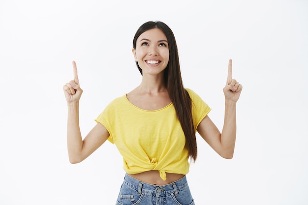 Charming carefree adult woman with long beatiful hair in yellow trendy t-shirt raising hands pointing and gazing upwards