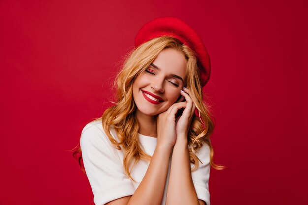 Charming caucasian girl with wavy hairstyle laughing on red wall.  cheerful french female model in beret.