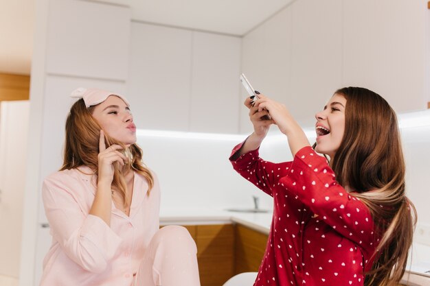 Charming curly girl posing with kissing face expression. Laughing european woman in red shirt using phone for photo of her lovely sister.