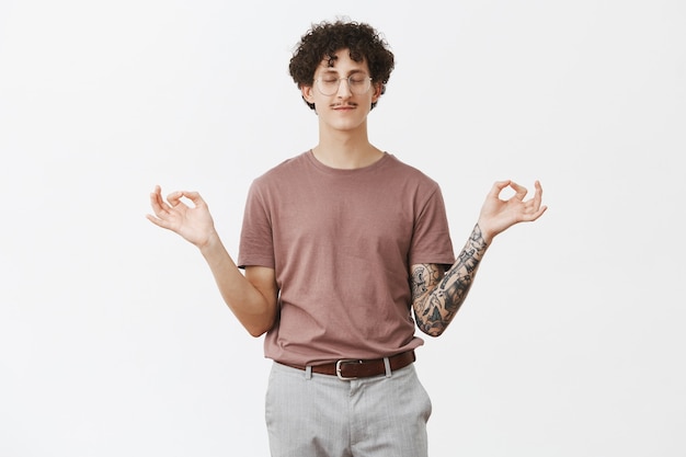 Free photo charming curly-haired guy with funny moustache standing in lotus pose trying find peace and comfort closing eyes and smiling with calm and relaxed expression meditating over gray wall