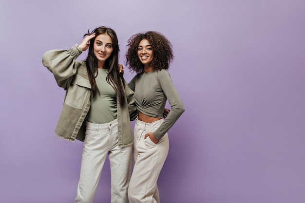 Free photo charming curly haired woman in olive top and white trousers smiling and hugging with stylish girl in jacket on lilac wall