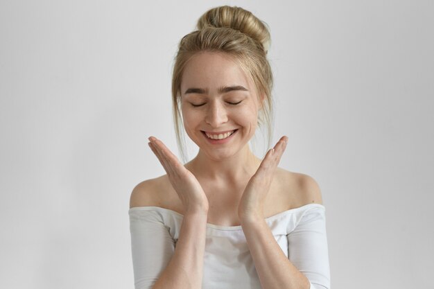 Charming European girl with hair knot closing eyes and smiling broadly, expressing joy and positive emotions, holding hands in front of her, receiving present, marriage proposal or good news