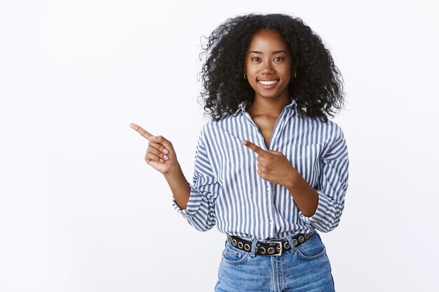 Charming friendly-looking smiling african-american assistant help you make choice grinning pleasant pointing left index finger promoting copy space product, standing joyfully upbeat white wall