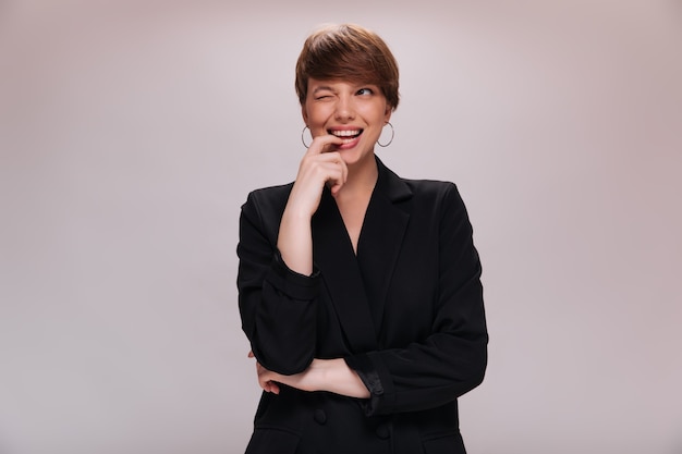 Charming girl in black jacket winking on isolated background. Short-haired woman in dark outfit smiles on white backdrop