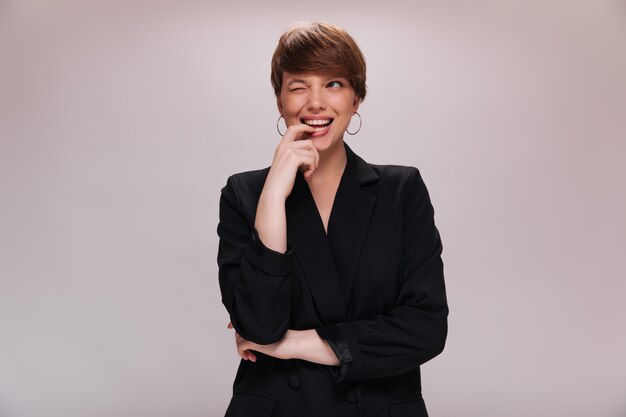 Charming girl in black jacket winking on isolated background. Short-haired woman in dark outfit smiles on white backdrop