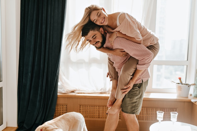 Charming girl blonde jumps on back to her boyfriend. Snapshot of lovers in great mood embracing.