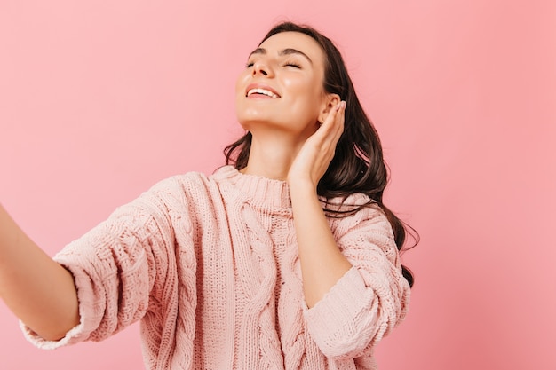 Charming girl smiles with closed eyes. Lady in beautiful sweater takes selfie in pink studio.