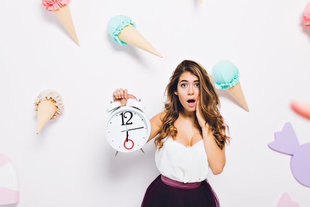 Free photo charming girl wearing luxury dress posing with surprised face expression on cute wall with sweet decor. portrait of worried young woman with shiny hair holding big clock.