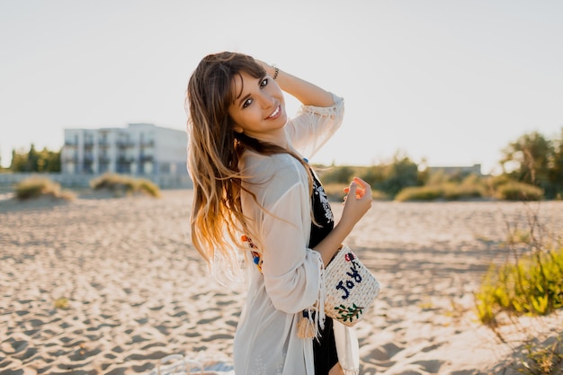 Free photo charming girl with wavy  brunette  hairs, dressed in  white boho cover up, walking on sunny summer beach.  traveling and vacation concept.