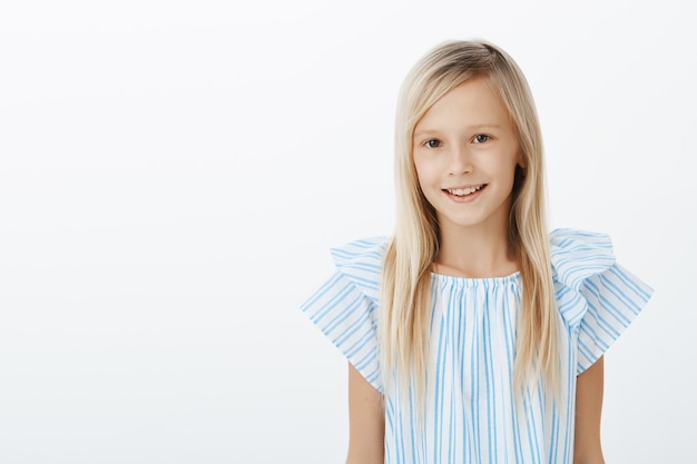 Charming happy young girl introducing herself to new classmates. Portrait of pleased carefree adorable daughter with blond hair in blue blouse, standing casually over gray wall and smiling friendly