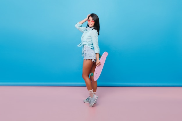 Charming hispanic woman with straight black hair standing on tip-toe and holding pink skateboard. Indoor photo of pretty asian female model in sunglasses wears sport shoes and denim shorts.