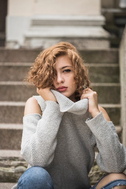 Charming lady holding sweater's collar and sitting on steps 