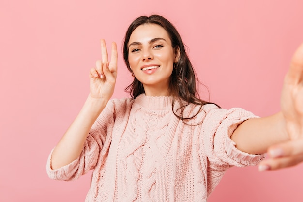 Charming lady in knitted dress makes selfie on pink background. Dark-haired woman in high spirits showing sign of peace.