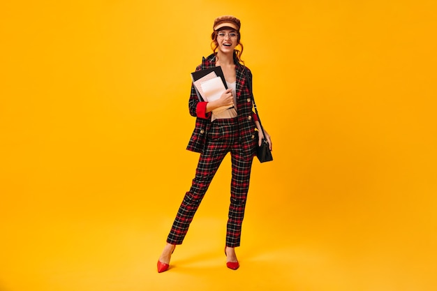 Free photo charming lady in plaid suit and cap poses with notebooks on orange wall