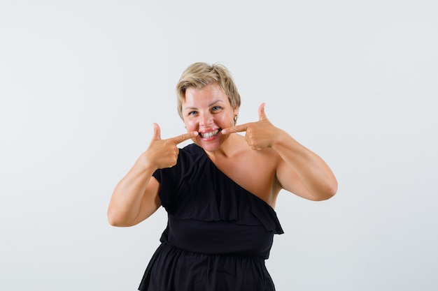 Charming lady pointing at her teeth in black blouse and looking satisfied. front view.