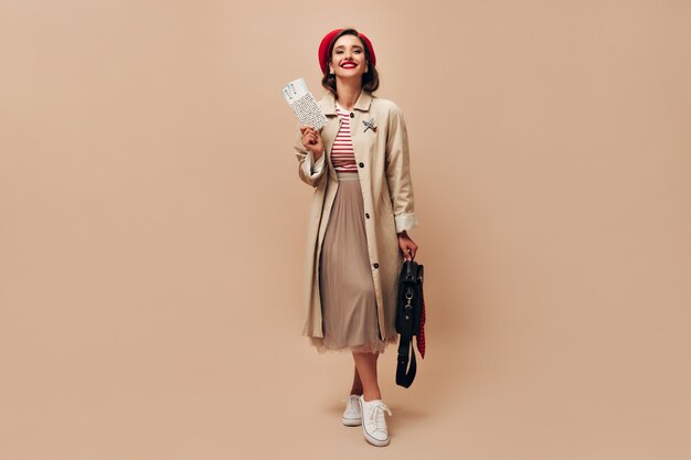 Charming lady in red hat and beige trench holds tickets. Beautiful woman in stylish long coat and in striped sweater with black bag posing.