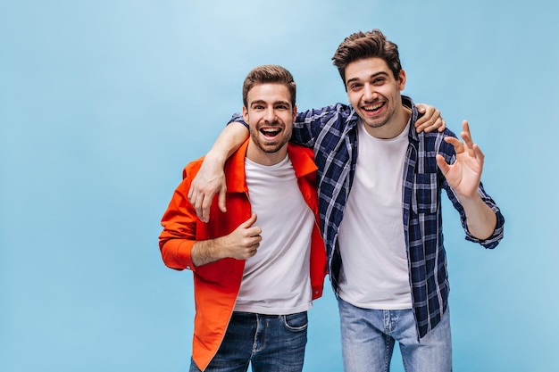 Charming man in orange jacket shows thumb up Brunet guy in checkered shirt and his friend smile on blue background