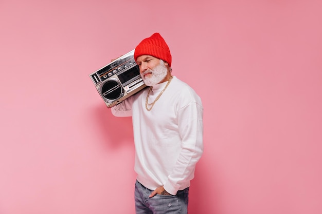 Charming man in stylish hat posing with record player on pink background Grey bearded adult guy in orange hat with gold chains smiling