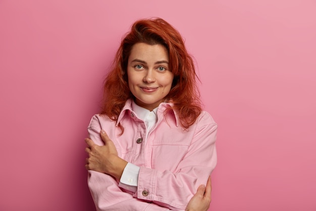 Free Photo charming natural woman embraces herself, has high self esteem, wears fashionable denim pink jacket, dimples on cheeks, foxy hair