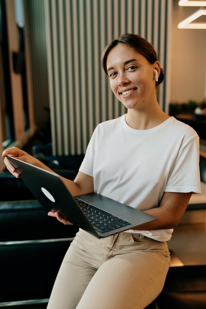 Charming pretty lady with wonderful smile and dark collected hair dressed white tshirt and beige pants is working at office with laptop