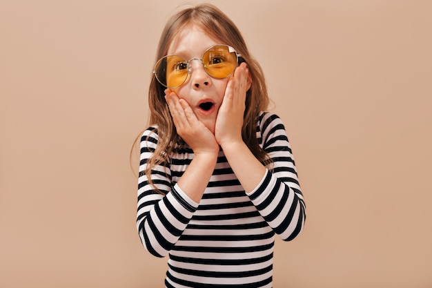 Free Photo charming pretty little 6 years old girl with light hair wearing stripped shirt posing with open mouth and holds hands on the check