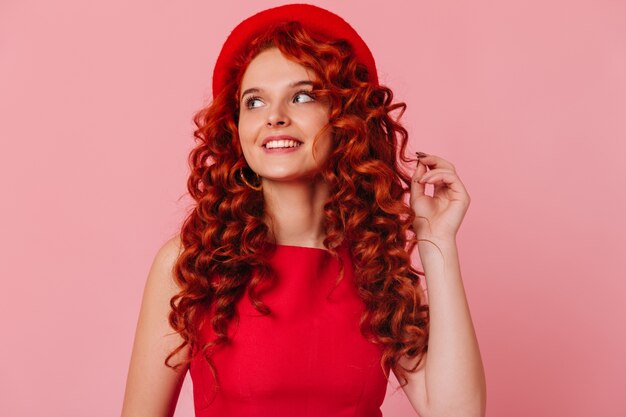 Charming red-haired girl with smile looks away. Woman in hat and red top touches hair.