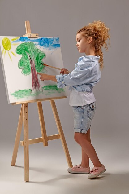 Charming school girl whith a curly blond hair, wearing in a blue shirt, white t-shirt and blue jeans shorts is painting with a watercolor brush on an easel, standing on a gray background.