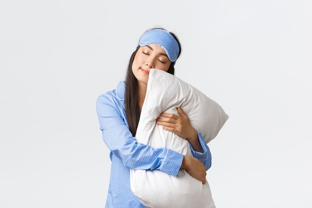 Charming sleeping asian girl in eyemask and pyjama, hugging pillow and lying on it with closed eyes, dreaming, having night sleep, posing over white background dreamy. Copy space