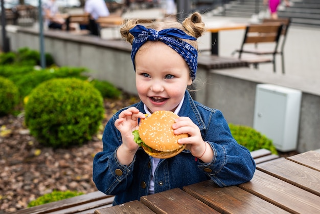 A charming smiling little girle is holding a hamburger in the open air on a sunny day.