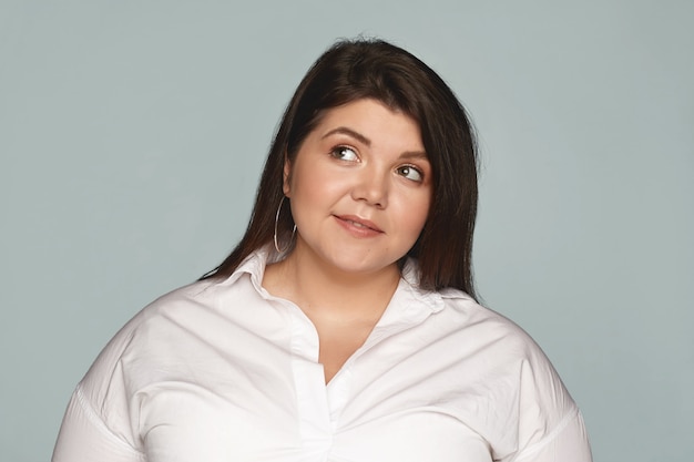 Charming stylish plus size young dark haired female looking up with thoughtful facial expression, daydreaming, thinking of something pleasant. Fashionable chubby woman in white shirt posing