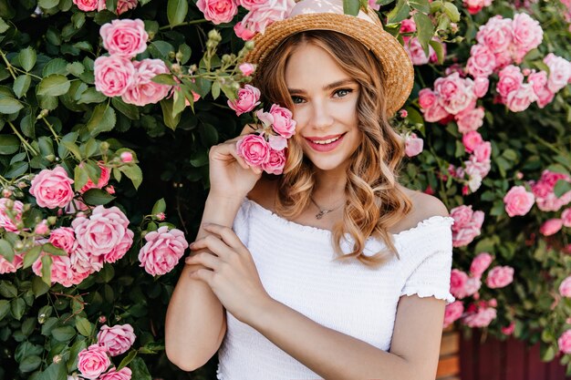 Charming white female model standing in front of pink flowers. Outdoor portrait of joyful girl in trendy hat spending time in garden.