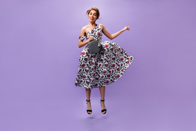 Charming woman in floral outfit jumping on purple background. Attractive stylish girl in colorful trendy dress smiling with handbag.