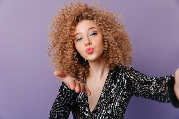 Free photo charming woman in great mood dressed in black top with silver ornament blows kiss and takes selfie on isolated space.