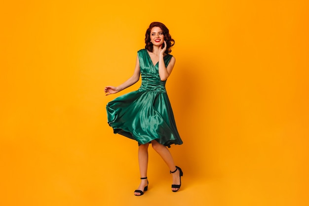 Free photo charming woman in green dress smiling and looking away. full length view of gorgeous curly girl dancing on yellow space.