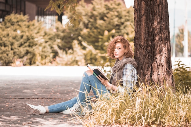 Free photo charming woman leaning on tree and holding book in park