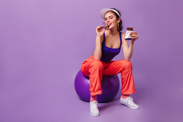 Charming woman in orange sweatpants and purple top is sitting on fitball and eating chocolate