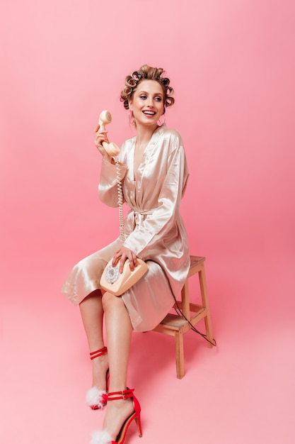 Free photo charming woman in pink robe smiles, sits on chair and holds phone on isolated wall