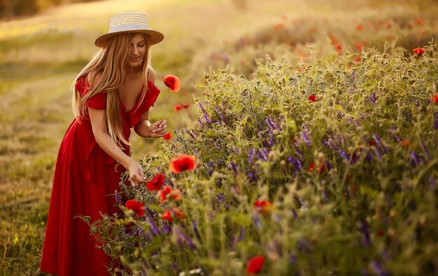 Charming woman walks across a green field with poppies in the rays of evening sun