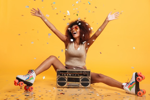 Free Photo charming young african woman in retro wear and roller scates throwing confetti while sitting with boombox