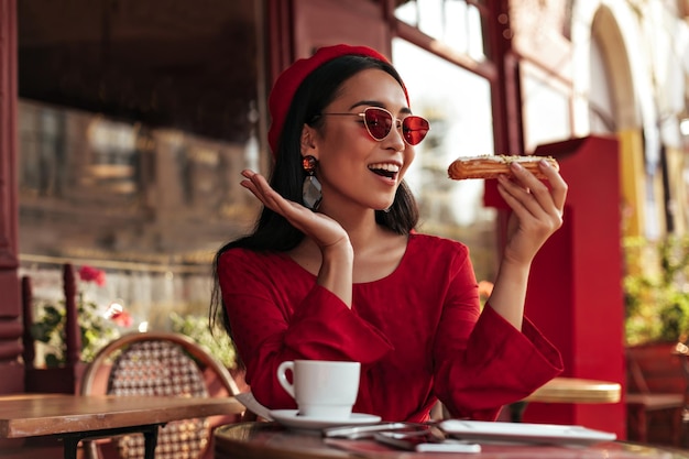 Charming young brunette woman in red beret bright colorful sunglasses and stylish dress looks at tasty eclair abc sits in cozy street cafe