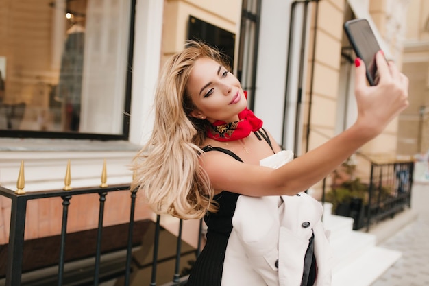 Charming young lady with nude make-up having fun outside and making selfie in beautiful dress. Adorable woman with long blonde hair taking photo of herself and smiling while walking around city.