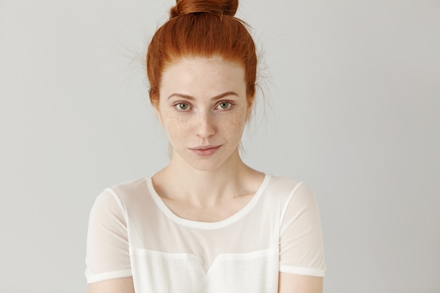 charming young redhead woman with hair bun standing against white blank wall