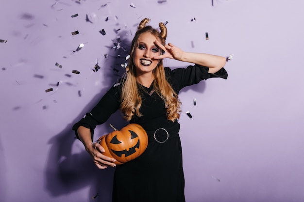 Charming young woman in black dress enjoying halloween carnival.  photo of smiling vampire girl holding orange pumpkin.