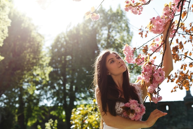 Free photo charming young woman in pink dress poses before a sakura tree full of pink flowers