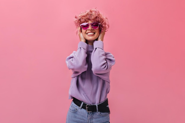 Free photo charming young woman in purple sweater and jeans smiles sincerely pinkhaired girl in fuchsia sunglasses poses on isolated background