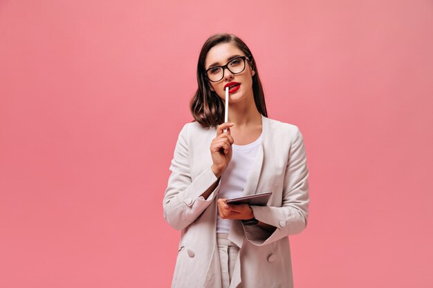 Charming young woman with dark hair in classic style suit and eyeglasses holds computer tablet on isolated pink background.