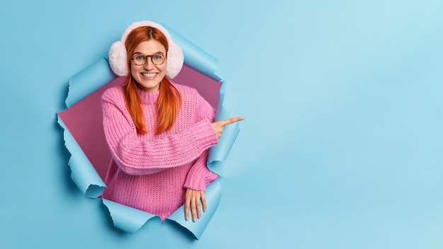 Free Photo cheeful lovely european woman with natural ginger hair wears warm earmuffs and knitted sweater pointing at copy space, breaks through paper hole.